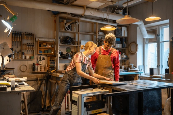 Two woodworkers, a woman and a man, standing by a table saw in a brightly lit, organized woodworking studio.