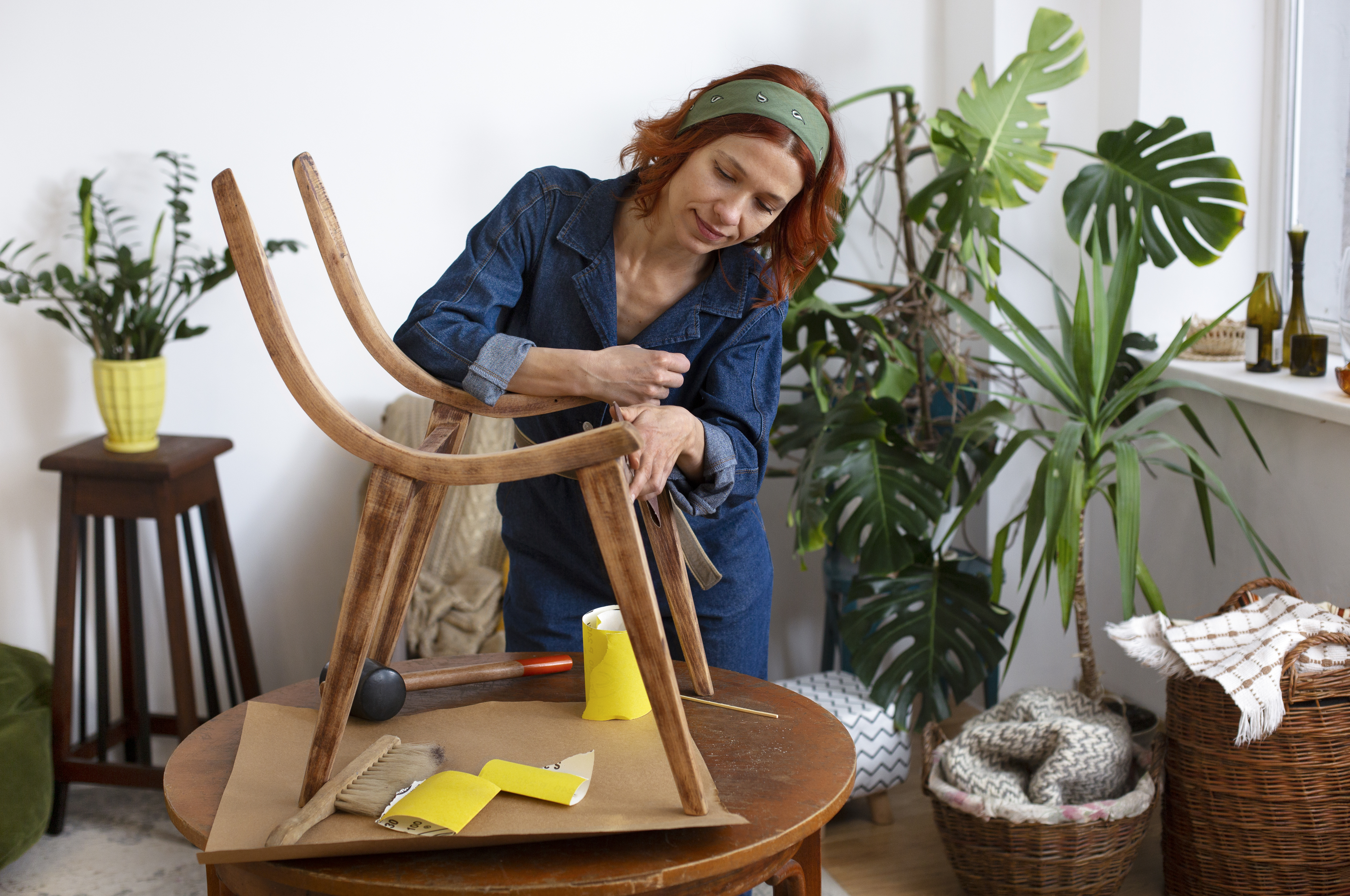 Woman sanding and repairing a wooden chair in a cozy room filled with indoor plants and natural light.