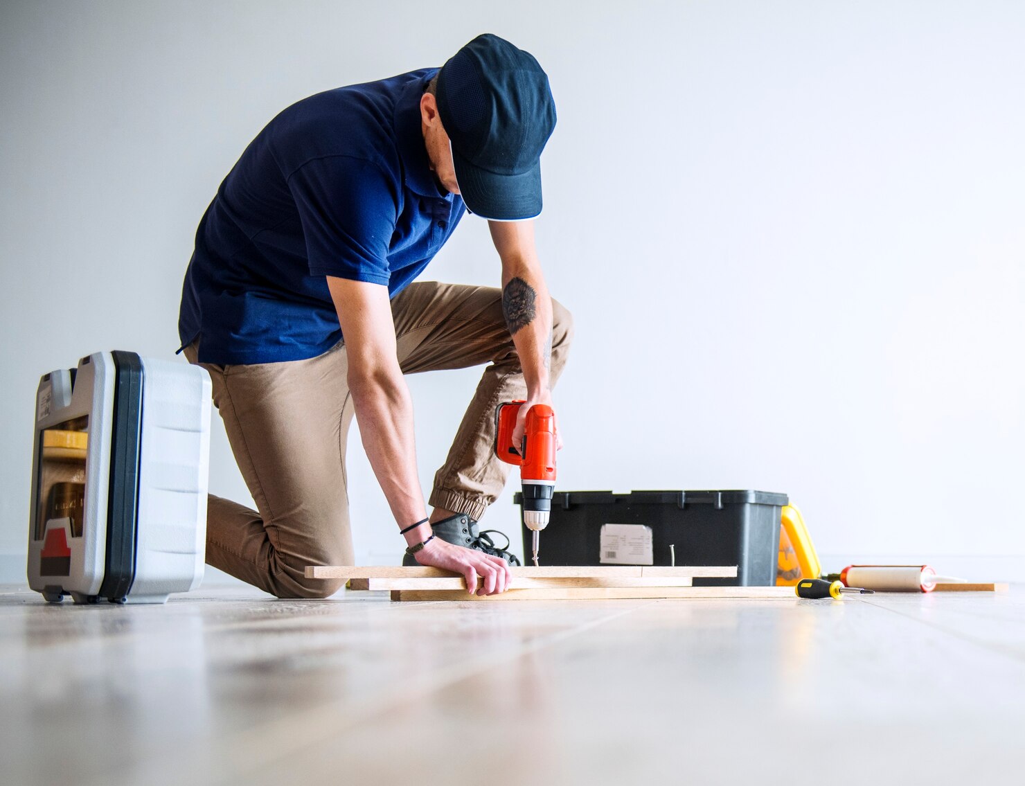Person using a power drill on wooden planks while kneeling on the floor, surrounded by tools and toolboxes.