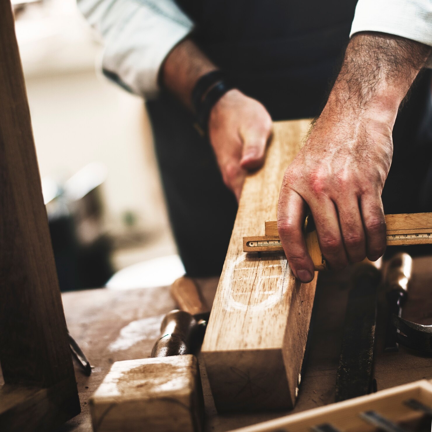 Close-up of a person measuring a wooden plank with a ruler on a workbench surrounded by woodworking tools.