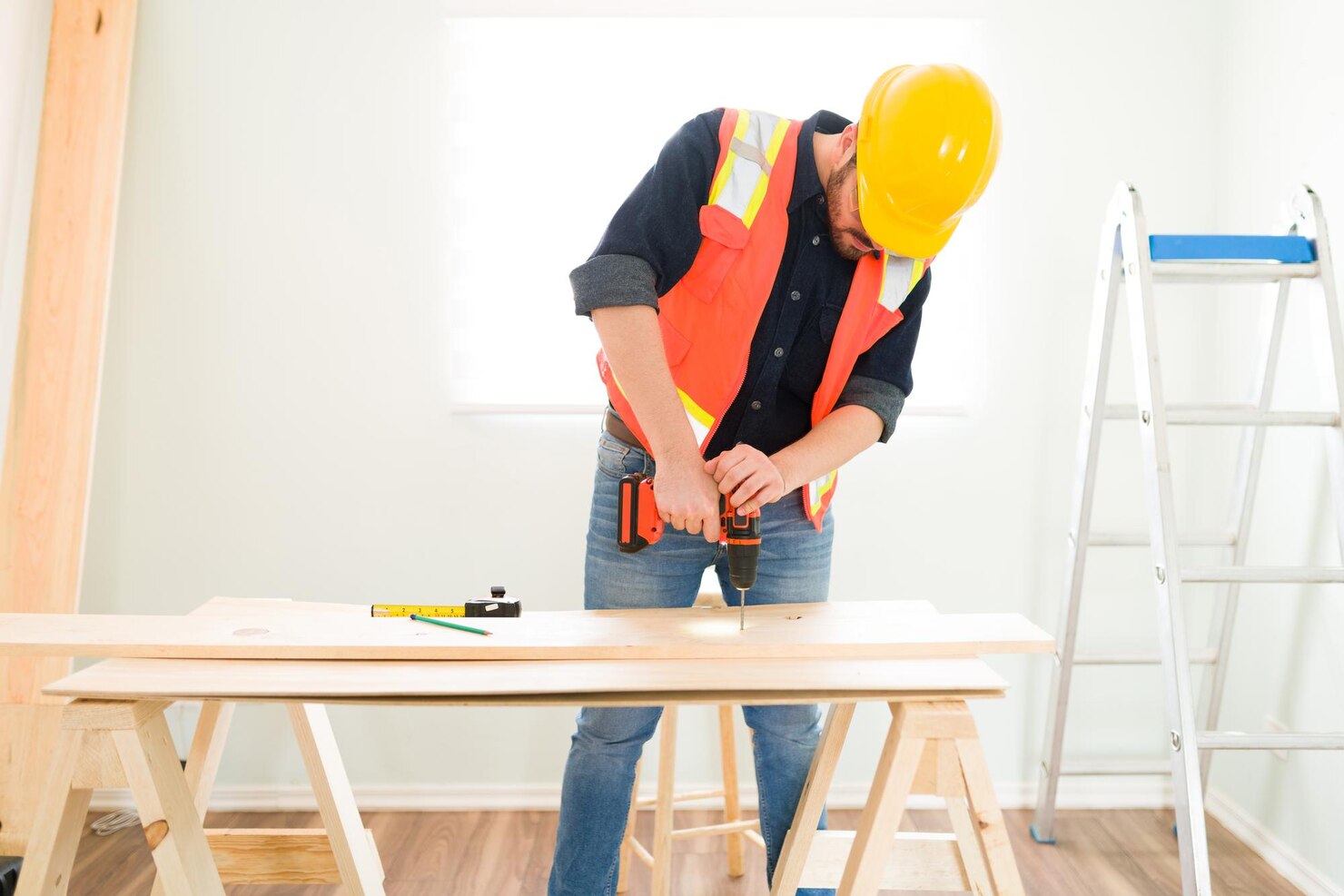 Construction worker wearing a safety vest and hard hat using an electric drill on a wooden board, with a ladder and measuring tools nearby.