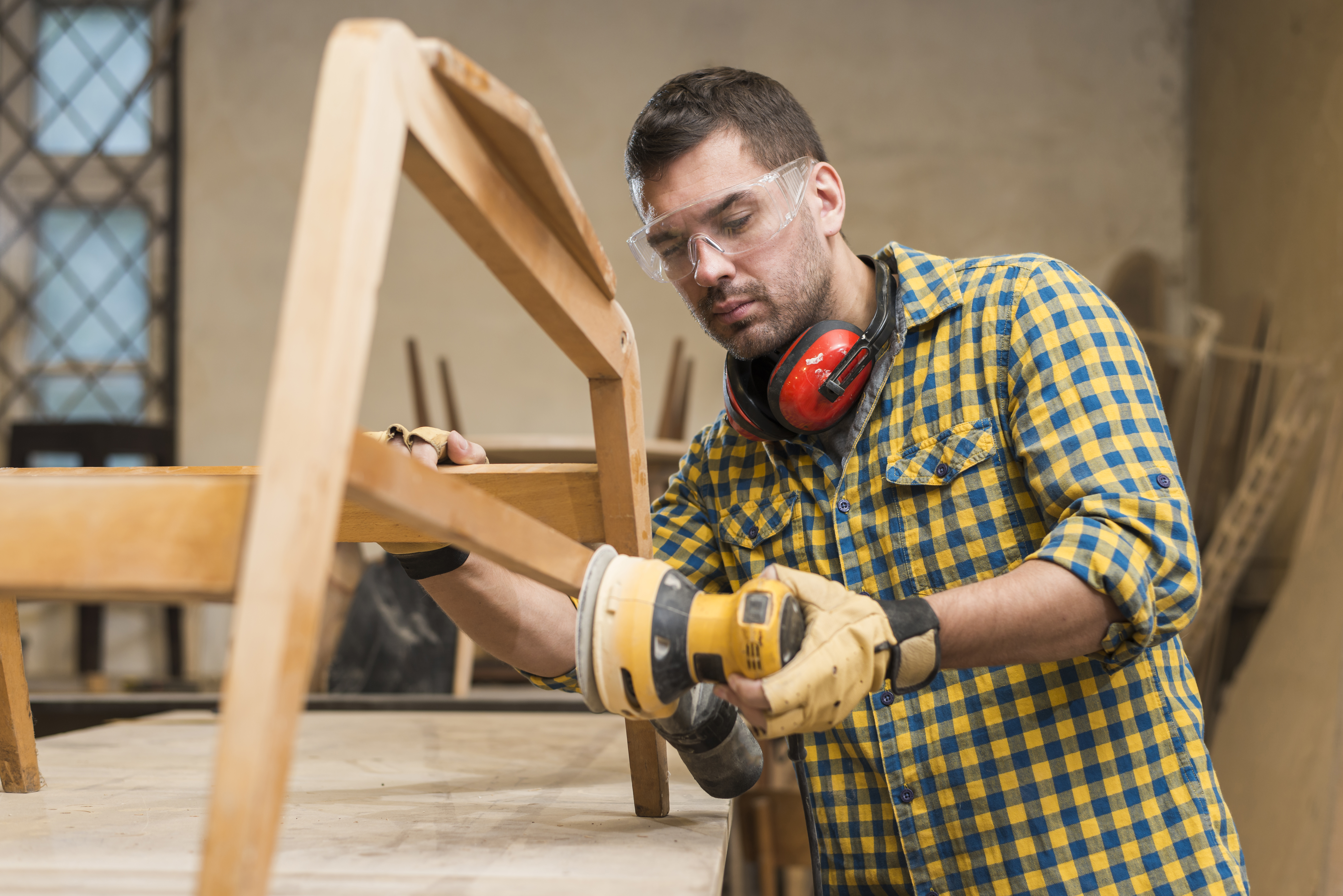 Carpenter wearing safety goggles and gloves sanding a wooden chair with an electric sander in a workshop.