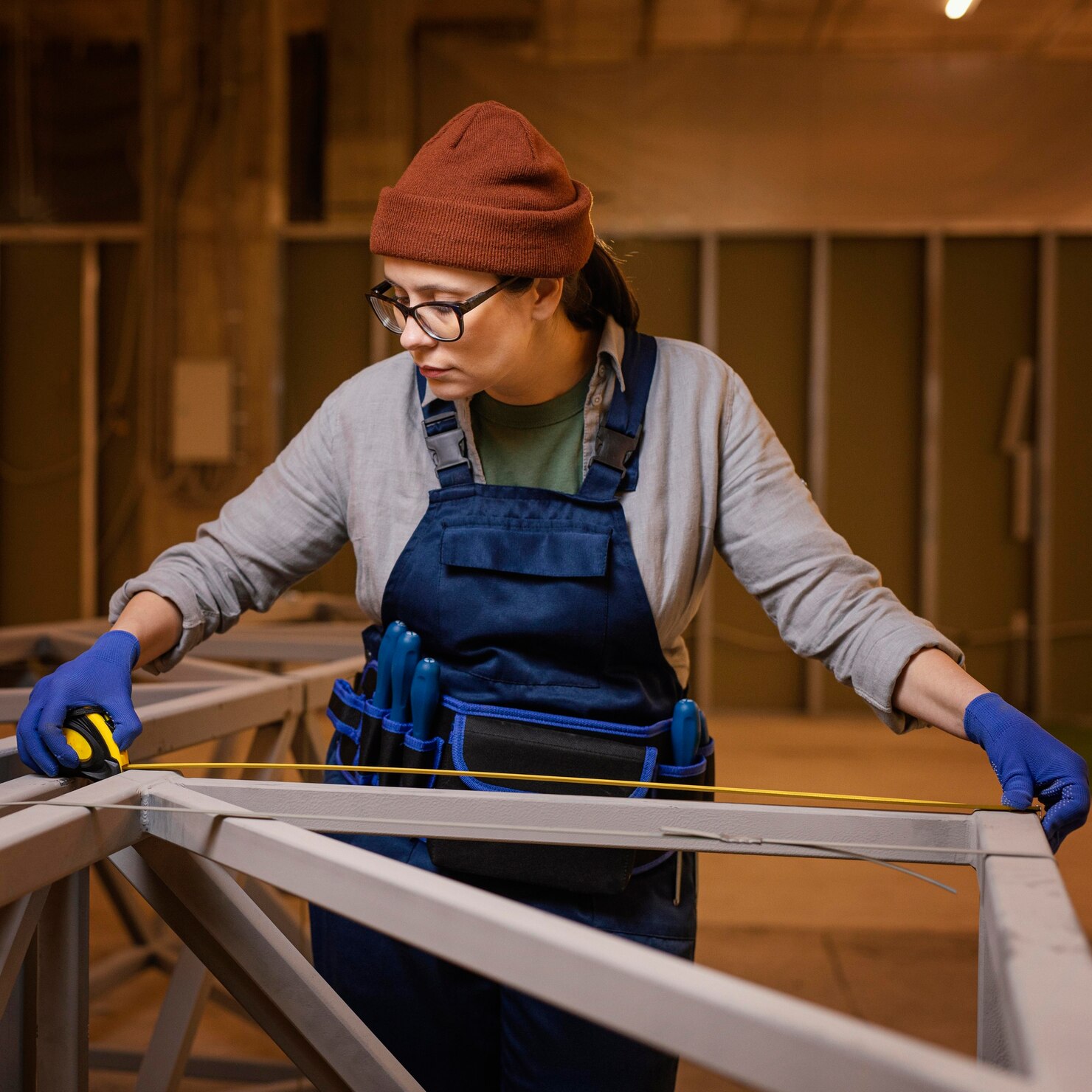 Person wearing a beanie, glasses, and work overalls using a tape measure to take precise measurements of a metal frame in a workshop.